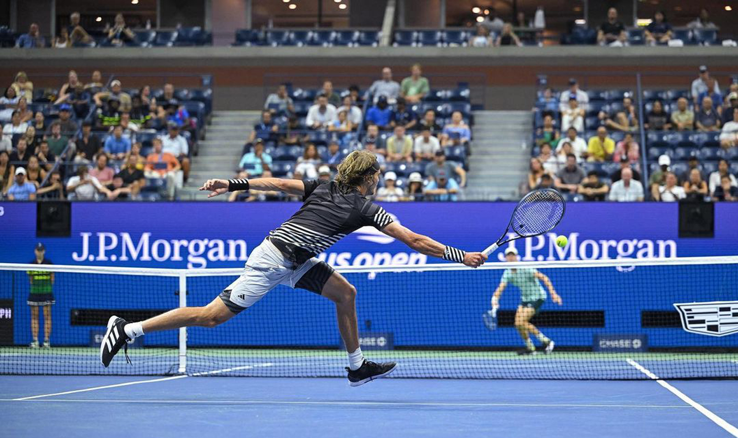 Александр Зверев в прыжке отбивает теннисный мяч ракеткой на корте US Open на фоне трибун с болельщиками
