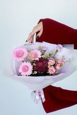 Букет «Для булочки» в стиле нежной флористики: розовые герберы, кустовые розы, сиреневые эустомы и бордовый шаровидный хризантем, которые держит девушка в бордовом свитере