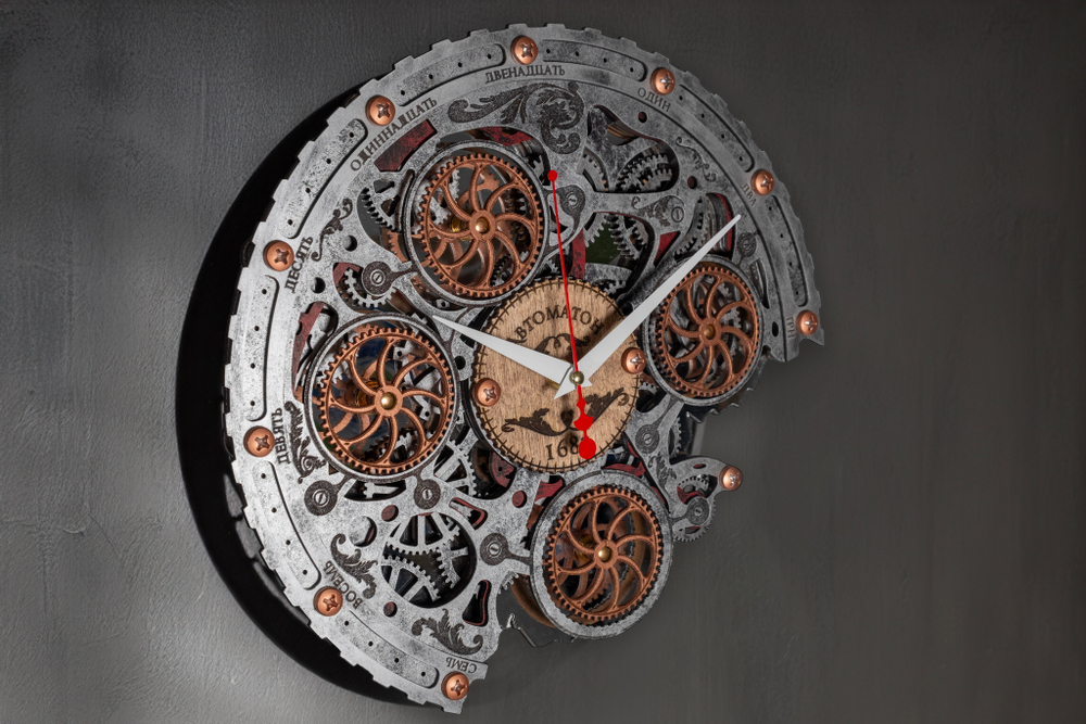 Настенные часы WOODandROOT Automaton Bite 1682 на серой стене с мягким светом, подчёркивающим открытый механизм и вращающиеся шестерёнки